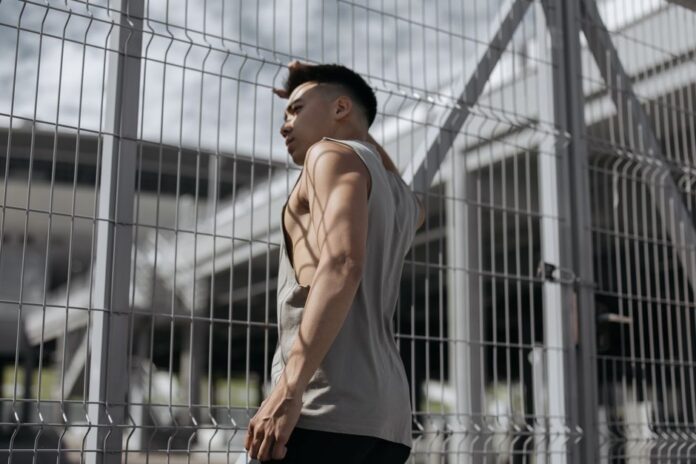 Young athletic man leaning against a metal fence outdoors, showcasing muscles and modern fashion.